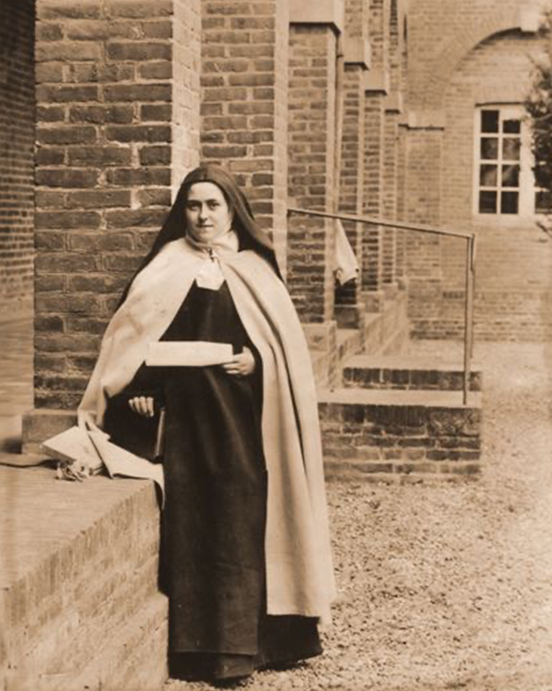 St. Therese in Carmelite habit leaning against a brick column with letter in hand.